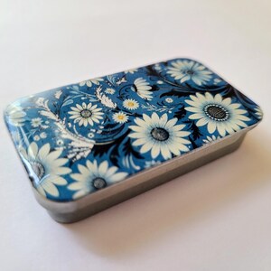 May include: A small, rectangular tin with a floral design. The tin is blue with white flowers and black accents.