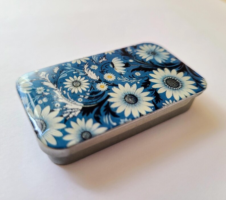 May include: A small, rectangular tin with a floral design. The tin is blue with white flowers and black accents.