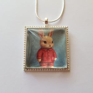 May include: A silver necklace with a square glass pendant featuring a cartoon bunny wearing a red shirt and pants. The bunny has brown fur and large brown eyes.