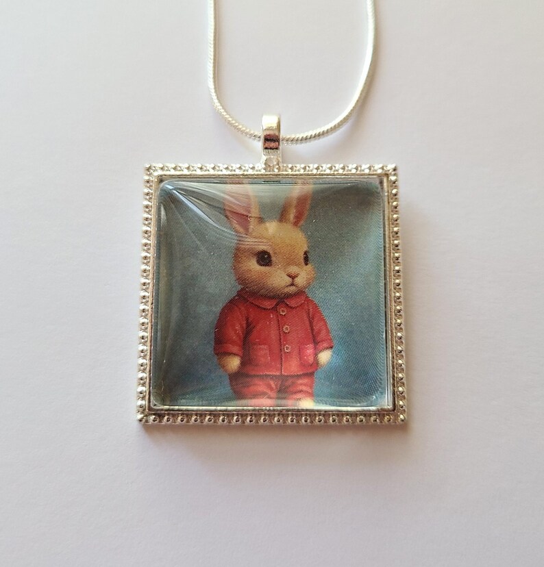 May include: A silver necklace with a square glass pendant featuring a cartoon bunny wearing a red shirt and pants. The bunny has brown fur and large brown eyes.