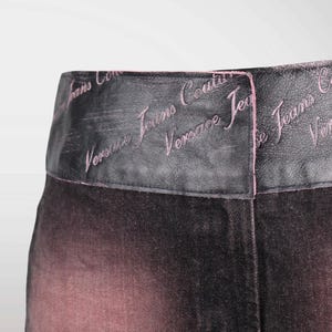 May include: Close-up of a pair of black and pink denim pants. The waistband is black with the words "Versace Jeans Couture" embroidered in pink. The denim fades from black to pink.