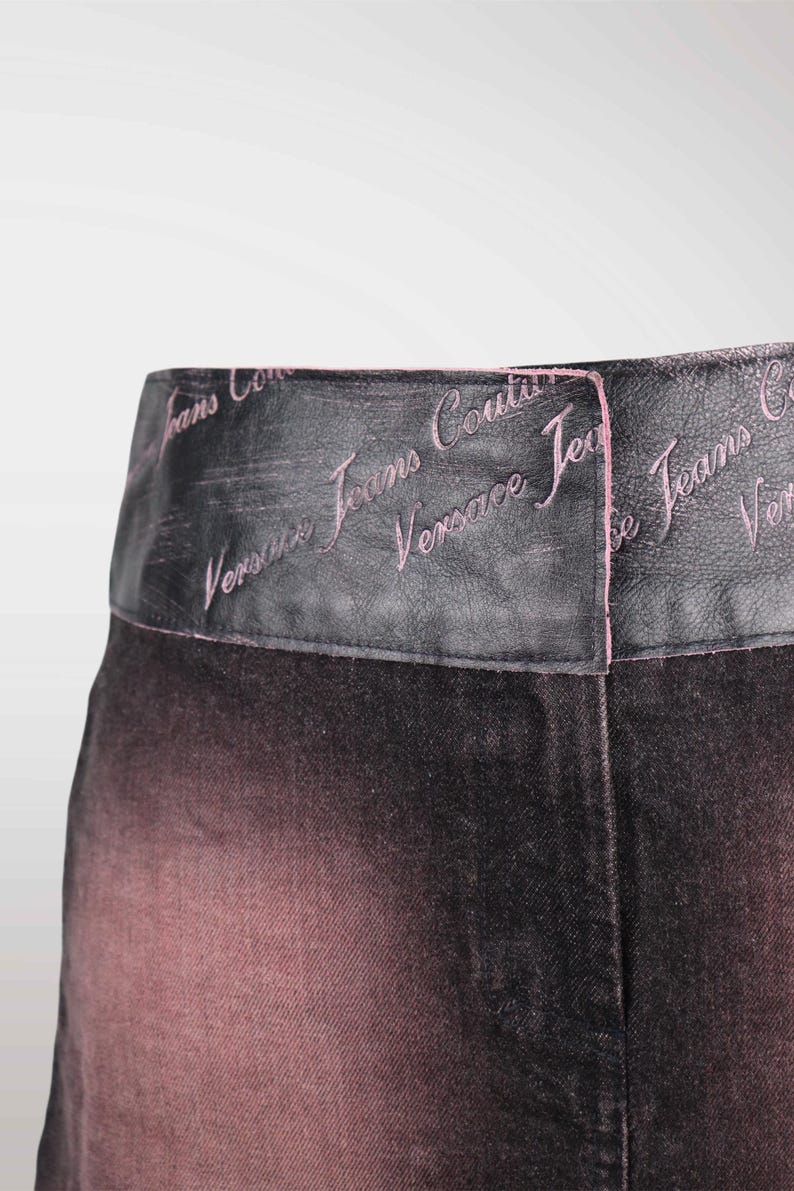 May include: Close-up of a pair of black and pink denim pants. The waistband is black with the words "Versace Jeans Couture" embroidered in pink. The denim fades from black to pink.