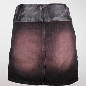 May include: A dark denim skirt with a faded pink and black gradient. The skirt features a black leather waistband with the words "Versace Jeans Couture" embroidered in pink.