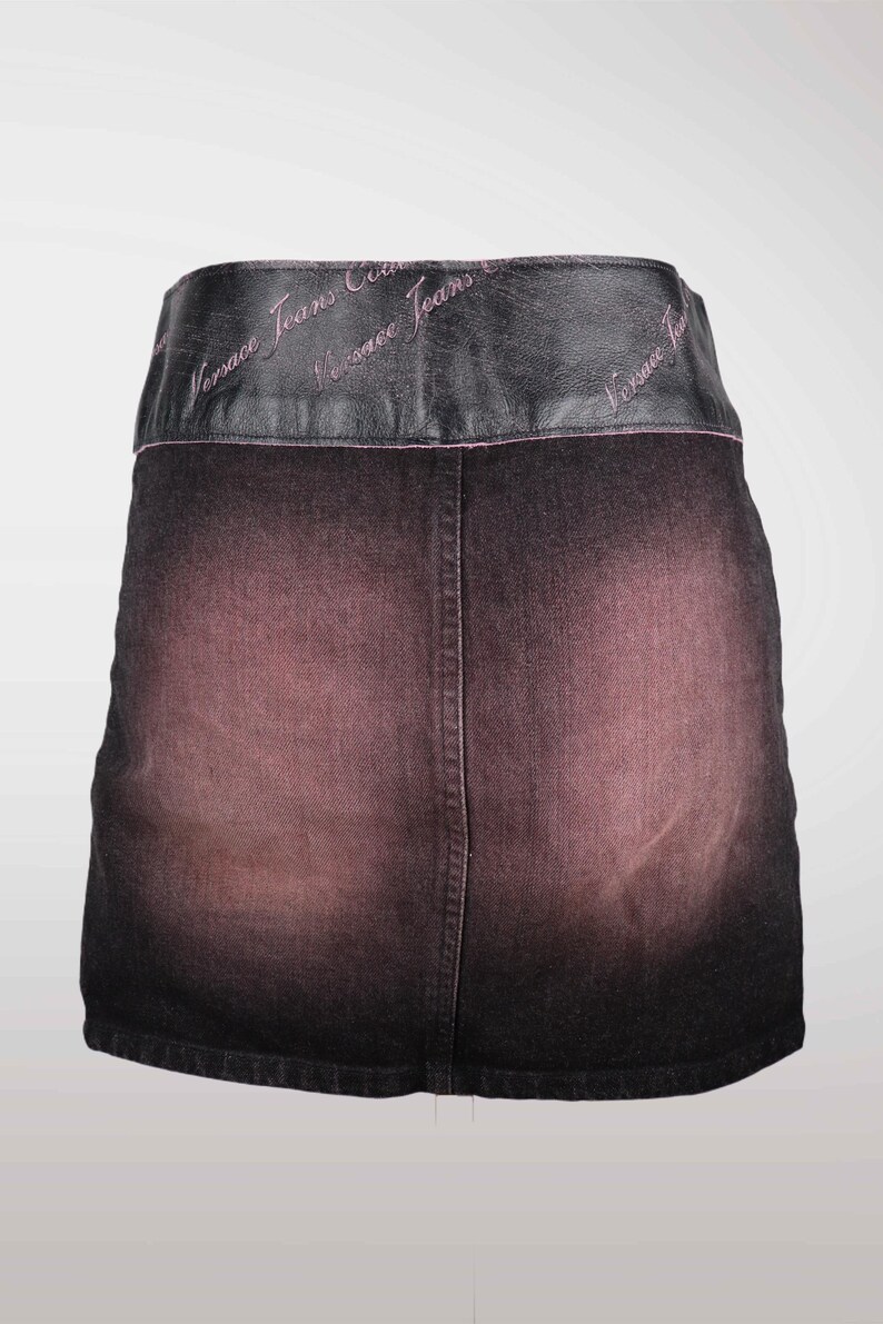 May include: A dark denim skirt with a faded pink and black gradient. The skirt features a black leather waistband with the words "Versace Jeans Couture" embroidered in pink.