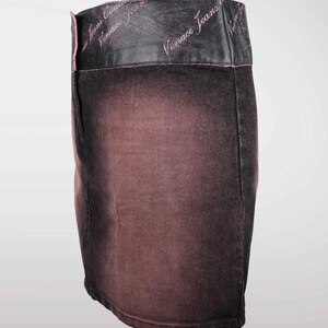 May include: A denim mini skirt with a faded, ombre effect in shades of brown and black. The waistband is black leather with the words "Versace Jeans" in pink script. A stylish, fashionable skirt.