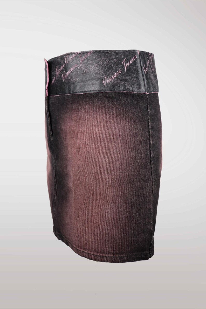 May include: A denim mini skirt with a faded, ombre effect in shades of brown and black. The waistband is black leather with the words "Versace Jeans" in pink script. A stylish, fashionable skirt.