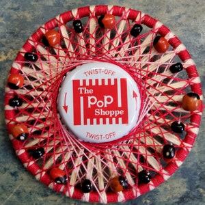May include: A red, white, and black string art piece with a red bottle cap in the center. The bottle cap has the text "The Pop Shoppe" on it. The string art is made with red, white, and black string and has red and black beads around the edge.