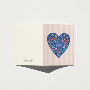 Puede incluir: Tarjeta de felicitaci&oacute;n con un dise&ntilde;o de coraz&oacute;n azul con flores y las palabras "Welcome Baby". El coraz&oacute;n est&aacute; sobre un fondo de rayas rosas y blancas. El lado izquierdo de la tarjeta es de color crema s&oacute;lido.