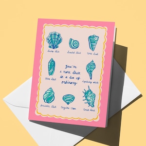May include: A pink greeting card with a light yellow border and illustrations of various seashells in teal. The card reads "You're a rare shell in a sea of ordinary" in blue script. The card is on top of a white envelope.