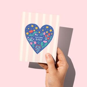 Puede incluir: Una tarjeta de felicitaci&oacute;n con un fondo a rayas rosas y blancas. La tarjeta presenta un coraz&oacute;n azul lleno de flores, estrellas y la frase "Welcome Baby". La tarjeta se sostiene sobre un fondo rosa.