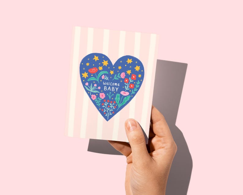 Puede incluir: Una tarjeta de felicitaci&oacute;n con un fondo a rayas rosas y blancas. La tarjeta presenta un coraz&oacute;n azul lleno de flores, estrellas y la frase "Welcome Baby". La tarjeta se sostiene sobre un fondo rosa.