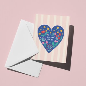 Puede incluir: Una tarjeta de felicitaci&oacute;n con un dise&ntilde;o de coraz&oacute;n azul lleno de flores y estrellas, con las palabras "welcome BABY". La tarjeta tiene un fondo a rayas rosas y blancas y va acompa&ntilde;ada de un sobre blanco. El fondo es rosa liso.