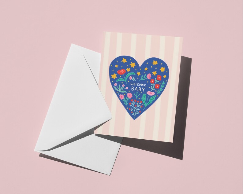 Puede incluir: Una tarjeta de felicitaci&oacute;n con un dise&ntilde;o de coraz&oacute;n azul lleno de flores y estrellas, con las palabras "welcome BABY". La tarjeta tiene un fondo a rayas rosas y blancas y va acompa&ntilde;ada de un sobre blanco. El fondo es rosa liso.