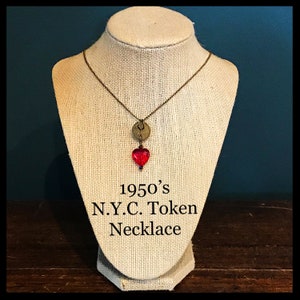 May include: A necklace with a red glass heart charm and a vintage 1950's New York City token charm. The necklace is on a brown fabric mannequin.