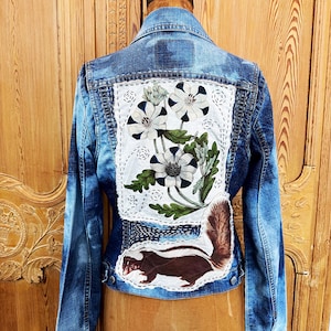 May include: Blue denim jacket with a floral and skunk patch design. The back of the jacket features a white patch with embroidered flowers and a skunk illustration. The jacket has long sleeves and a classic denim jacket style.