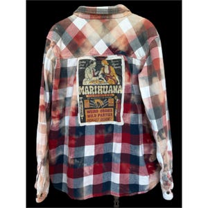 May include: Plaid flannel shirt in red, white, and brown tones with a distressed, bleached effect. The back features a rectangular patch with the word "Marihuana" and vintage-style illustrations and text.