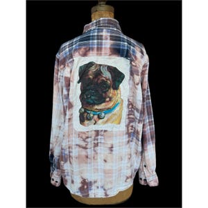 May include: A long-sleeved, button-down flannel shirt with a unique design. The shirt features a large, rectangular patch on the back showcasing a detailed pug illustration wearing a blue collar with bells. The shirt has a plaid pattern in shades of brown, blue, and white.