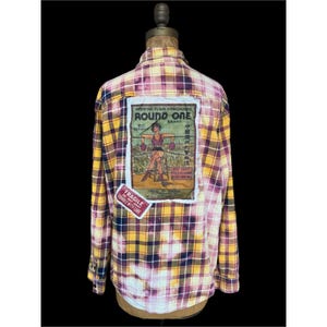 May include: A plaid button-up shirt in yellow, purple, and white, with a vintage graphic patch on the back that reads "ROUND ONE" and a "FRAGILE" sticker. The shirt is displayed on a mannequin.