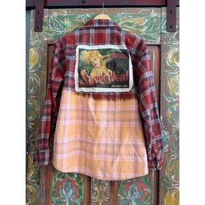 May include: A red and black plaid flannel shirt with a bleached orange and pink lower half. A rectangular patch on the back features a vintage-style image of a woman and the text "The Devil's Weed" and "For Adults Only".