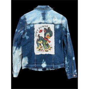 May include: A blue denim jacket with a light blue wash on the upper back and sleeves. The back features a white patch with a cartoon deer illustration and the words "SOFT POWER".
