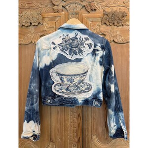 May include: A denim jacket with a unique design. The jacket features a light blue and white tie-dye effect. The back of the jacket is adorned with a floral patch and a teacup and saucer design in blue and white.