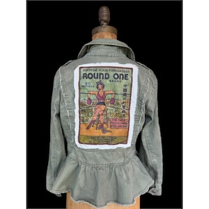 May include: An olive green jacket with a peplum waist and a large, rectangular patch on the back. The patch features a vintage-style illustration of a woman in boxing gloves, with text that reads "ROUND ONE" and "SUPERFINE FLASH FIRECRACKERS".