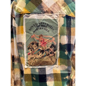 May include: A button-down shirt with a colorful plaid pattern in shades of yellow, green, and blue. A rectangular patch on the back features a vintage illustration of a woman riding a horse, with the text "LOVE IS WHAT MAKES THE RIDE WORTHWHILE."