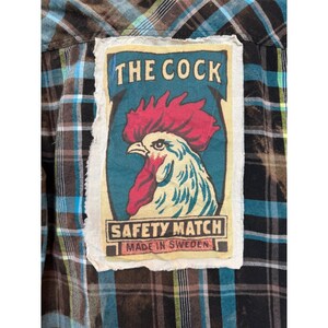 May include: A plaid shirt in shades of brown, blue, and black, featuring a rectangular patch with a rooster illustration. The patch reads "THE COCK SAFETY MATCH MADE IN SWEDEN" in a vintage style.