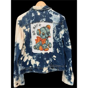 May include: A denim jacket with a blue and white tie-dye pattern. The back features a patch with a cartoon teddy bear and the words "BE NICE". The jacket has a classic button-up design.