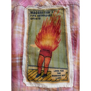 May include: Pink plaid shirt with a rectangular patch featuring a cartoon figure engulfed in flames. The patch reads "MACGREGOR'S FIRE RETARDANT SHORTS" and "LOOK FOR THE BURNING BOY LABEL". The figure is wearing red shorts and black shoes.