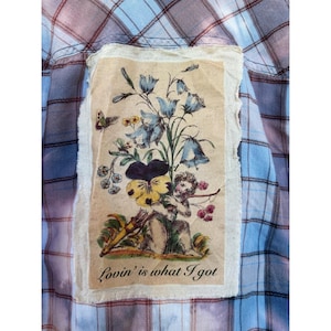 May include: A light blue and purple plaid shirt featuring a rectangular fabric patch with raw edges. The patch displays a printed illustration of a cherub holding a large pansy and bluebell flowers, with the text Lovin' is what I got below.