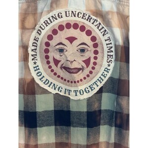 May include: A plaid flannel shirt in shades of brown, tan, and navy blue. A circular patch is sewn on the back, featuring a smiling sun face design with the text "MADE DURING UNCERTAIN TIMES* HOLDING IT TOGETHER".