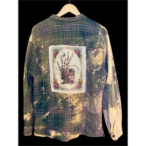May include: Green and purple plaid flannel shirt with a bleached design. A patch on the back features a floral design with a cat, lavender, and the word "Kindness". The shirt is hanging on a wooden hanger.