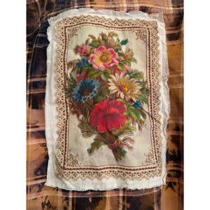 May include: A brown and black plaid flannel shirt with a floral patch. The patch features a vintage-style bouquet of red, pink, blue, and white flowers, framed by a decorative border and lace trim.