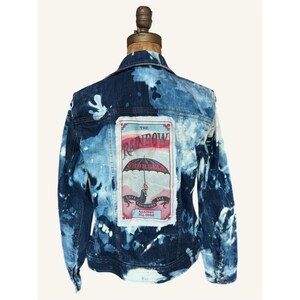 May include: A blue denim jacket with a bleached tie-dye pattern featuring a rectangular patch on the back. The patch displays an umbrella illustration with the text THE RAINBOW UMBRELLA and LOVE CONQUERS ALL AGAINST ALL ODDS.