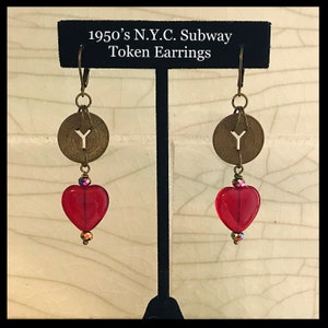 May include: A pair of earrings featuring vintage New York City subway tokens and red heart-shaped beads. The earrings are suspended from bronze-colored ear wires. The text "1950's N.Y.C. Subway Token Earrings" is displayed above the earrings.