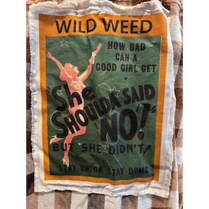 May include: A vintage-style fabric sign with the words "WILD WEED" at the top. The sign features a retro illustration of a woman and the text "She Shoulda Said NO!" in a green and orange color scheme.