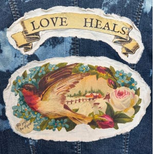May include: A denim jacket with two fabric patches. The top patch reads "LOVE HEALS" on a banner. The bottom patch features a bird, flowers, and a winter scene with the text "All joys be thine".