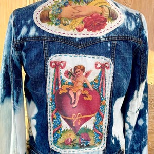 May include: A blue denim jacket with a bleached design. The back features two patches with vintage-style illustrations. One shows clasped hands and flowers, the other a cherub on a heart. The jacket has a classic collar and button closure.