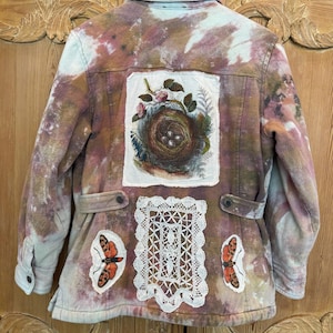 May include: A tie-dye denim jacket with a bird's nest patch, lace applique, and butterfly patches. The jacket features a button-down front, pockets, and a unique, artistic design. The color palette includes shades of brown, pink, and white.