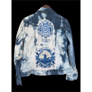 May include: A denim jacket with a blue and white tie-dye effect. The back features two patches with blue floral and windmill designs. The jacket has a classic collar, button closures, and long sleeves.