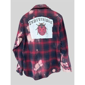 May include: Red and black plaid flannel shirt with a distressed, bleached design. The back features a white patch with the word "INDIVISIBLE" above a red ladybug illustration. The shirt has a button-down front and long sleeves.