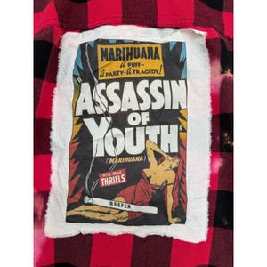 May include: A red and black plaid flannel shirt with a white patch featuring vintage-style text and graphics. The patch reads "Marihuana, Assassin of Youth" with a graphic of a person and a lit cigarette.