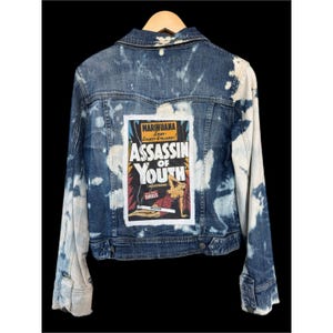 May include: A denim jacket with a bleach-splattered design. The back features a rectangular patch with the words "Marihuana" and "Assassin of Youth" in a vintage comic book style. The jacket has a classic collar and button closures.