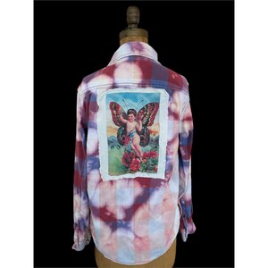 May include: A long-sleeved, button-down shirt with a tie-dye effect in shades of red, white, and blue. The back features a rectangular patch with a vintage illustration of a cherub with butterfly wings, surrounded by flowers.