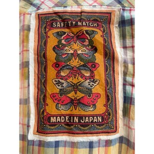 May include: A colorful plaid shirt with a rectangular patch featuring a butterfly design. The patch has the words "SAFETY MATCH" at the top and "MADE IN JAPAN" at the bottom. The butterflies are in shades of red, blue, and yellow.