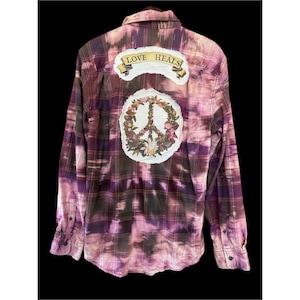 May include: A long-sleeve button-down shirt featuring a purple and dark plaid pattern with a bleached tie-dye effect. The back displays a white patch with a floral peace sign and a banner above it with the text LOVE HEALS in black letters.