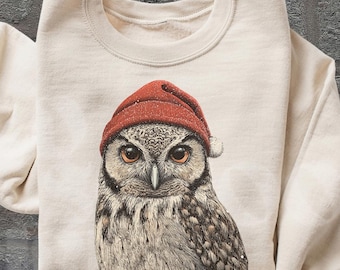 May include: Cream-colored sweatshirt featuring a detailed illustration of an owl wearing a red knit cap. The owl has brown and white feathers and large, expressive eyes. The sleeves are rolled up. The words "OLD SCHOOL CURIOS" are in the bottom right corner.