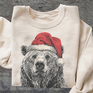 May include: Cream-colored sweatshirt featuring a detailed illustration of a bear wearing a red Santa hat. The bear's fur is depicted in shades of gray and black. The sweatshirt has long sleeves and a crew neck. The words "OLD SCHOOL CURIOS" are visible in the lower right corner.