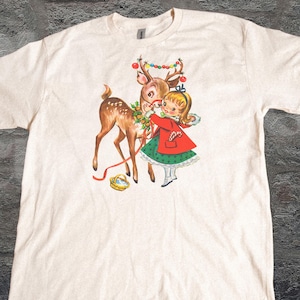 Può includere: T-shirt color crema con un'illustrazione vintage di una bambina con cappotto rosso e vestito verde, che decora un cervo con ornamenti natalizi. Sono visibili anche un cestino dorato e il testo "OLD SCHOOL CURIOS".